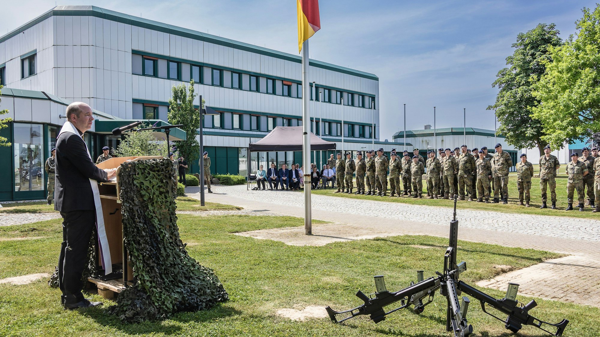 Vor dem Kasernengebäude spricht der katholische Militärpfarrer Lars Spohr zu den angetretenen Soldatinnen und Soldaten und Gästen. Vor ihm sind Sturmgewehre zusammengestellt.