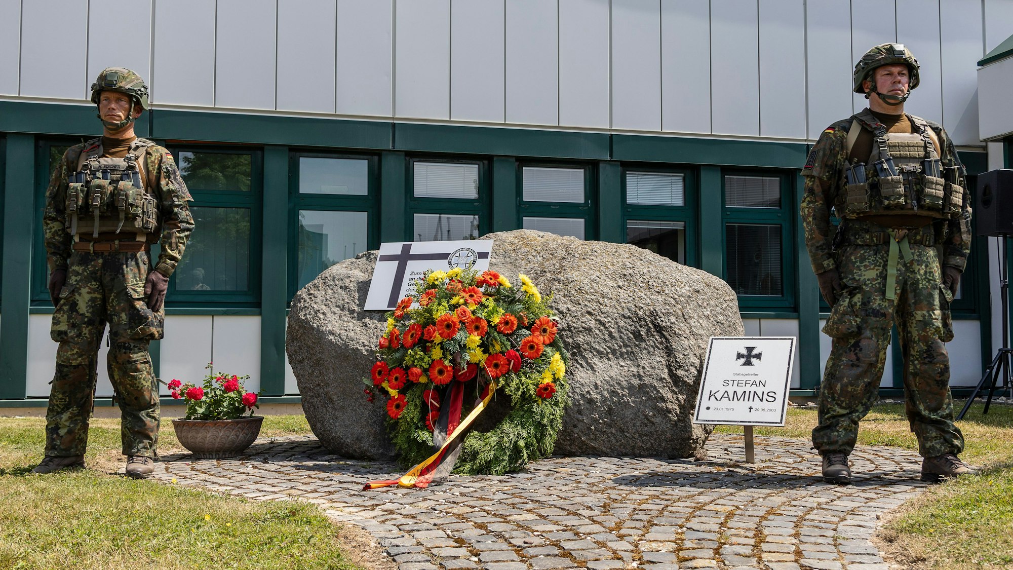 Zwei voll ausgerüstete Soldaten stehen neben einem Stein, an dem sie zuvor einen Kranz niedergelegt haben. Ein Schild weist auf den in Afghanistan gefallenen Stabsgefreiten Stefan Kamins hin.