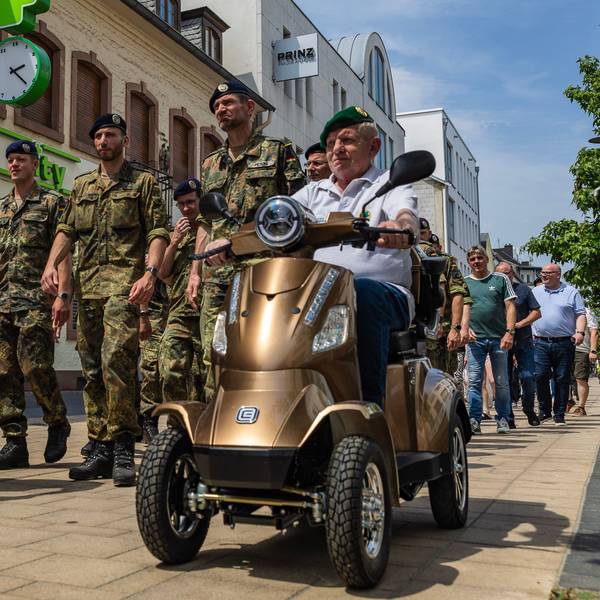 Soldaten in Feldanzügen marschieren auf der Neustraße. Vor ihnen ein Ex-Soldat mit Rollator und ein weiterer mit grünem Barett auf einem Elektromobil.