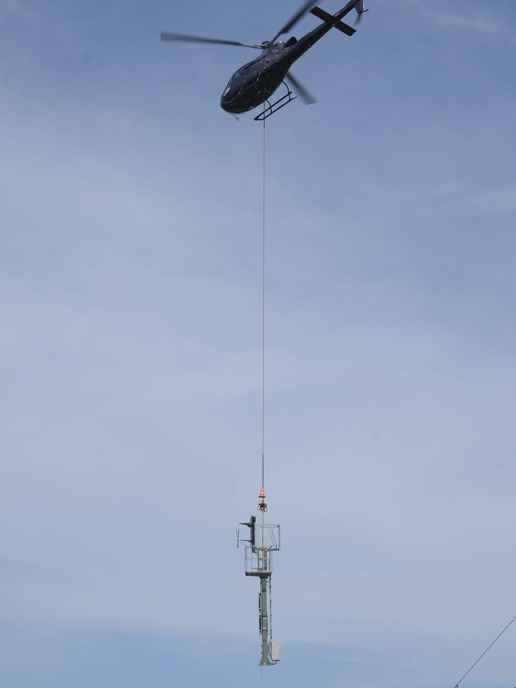 Ein Helikopter transportiert einen Mast an einem langen Stahlseil.