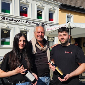 Zu sehen sind Margherita (l.) und Francesco Attrice (r.) mit jeweils einer Weinflasche in der Hand sowie in der Mitte ihr Vermieter Rolf Dieffendahl.