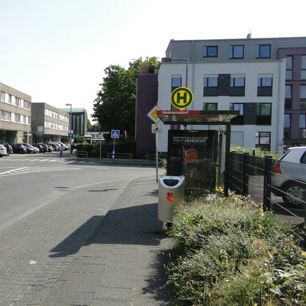 An einer Straße liegt eine Bushaltestelle, auf der Straßenseite gegenüber ist ein schmuckloser dreistöckiger Bau, der Haselnusshof, zu sehen.