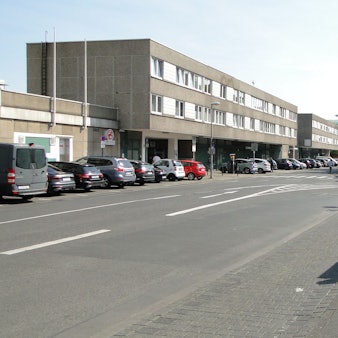 An einer Straße liegt eine Bushaltestelle, auf der Straßenseite gegenüber ist ein schmuckloser dreistöckiger Bau, der Haselnusshof, zu sehen.