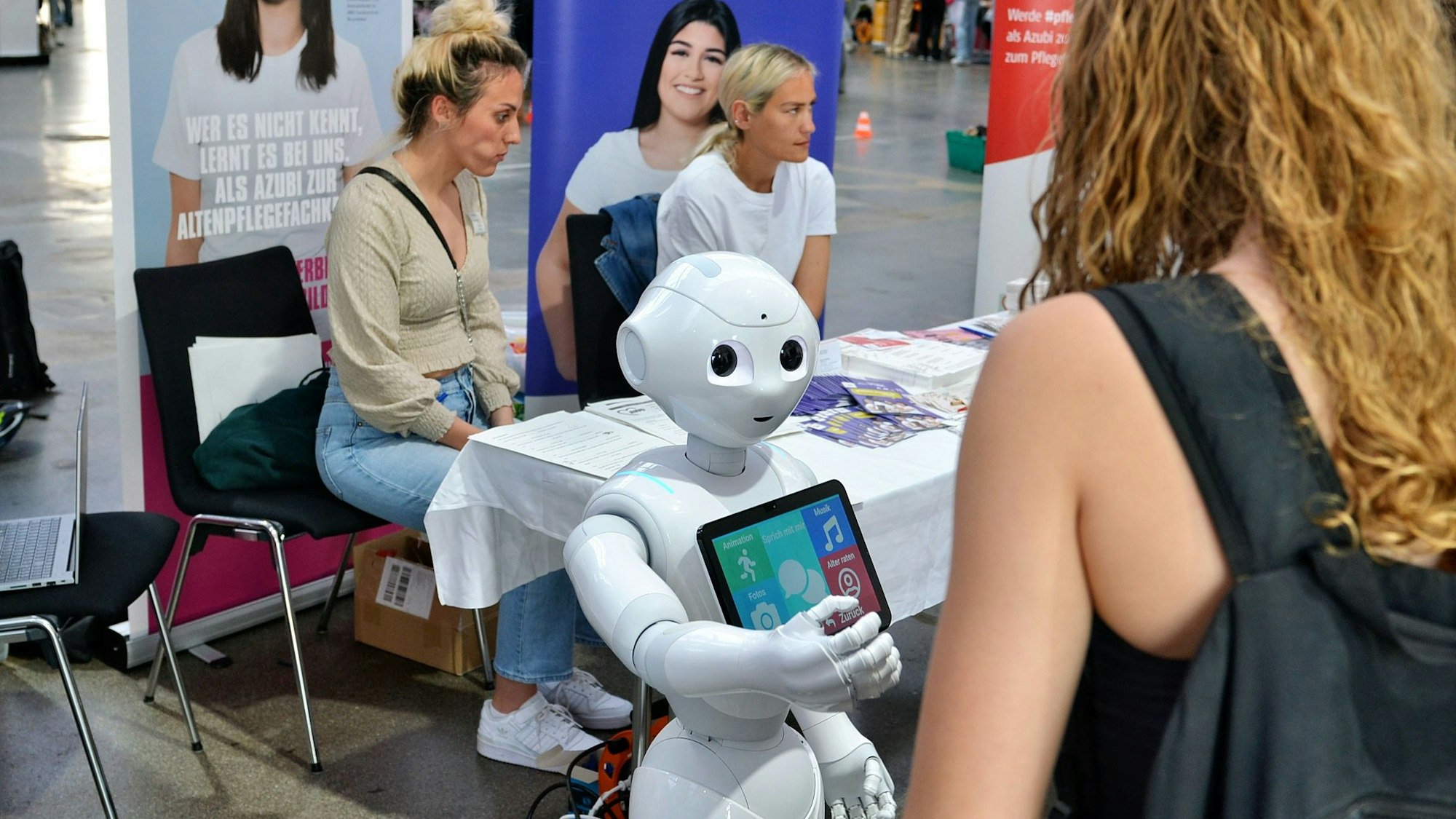 Das Bild zeigt den Roboter vor einem Infotisch, an dem zwei Frauen sitzen.