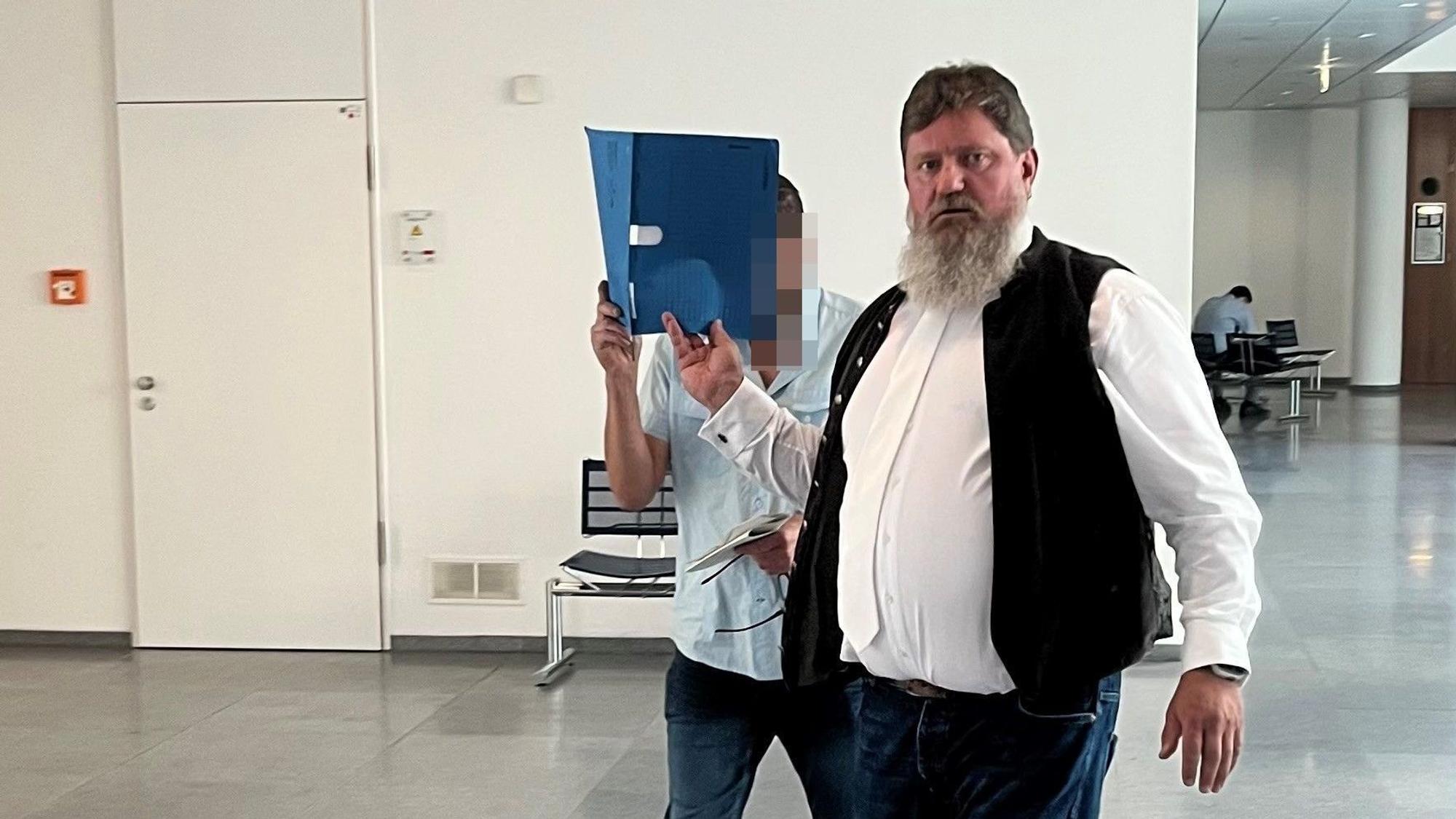Ein Mann mit Bart schaut in die Kamera. Ein zweiter Mann, Angeklagter in einem Prozess, hält sich einen blauen Aktenordner vors Gesicht.