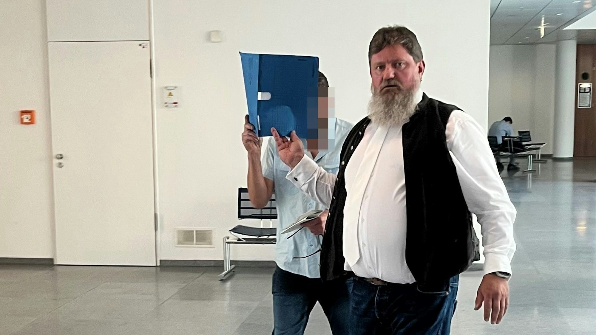 Ein Mann mit Bart schaut in die Kamera. Ein zweiter Mann, Angeklagter in einem Prozess, hält sich einen blauen Aktenordner vors Gesicht.