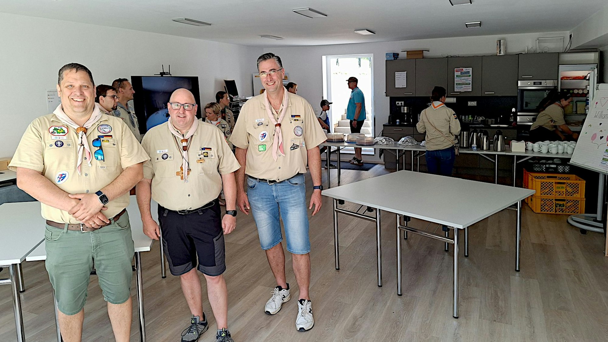 Das Bild zeigt drei Männer in Pfadfindermontur in einem Raum mit Tischen und Küchenzeile.