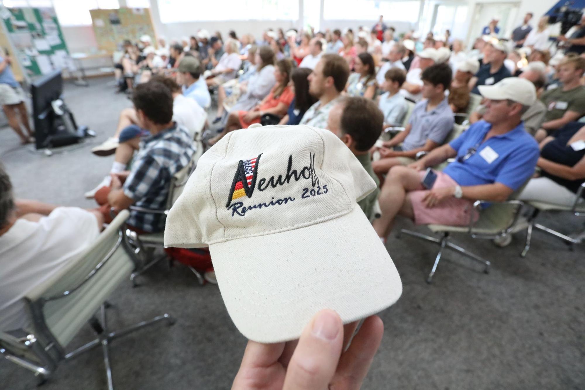 Eine Basecap mit der Aufschrift „Neuhoff – Reunion 2025“, dahinter viele Menschen, die auf Stühlen sitzen.