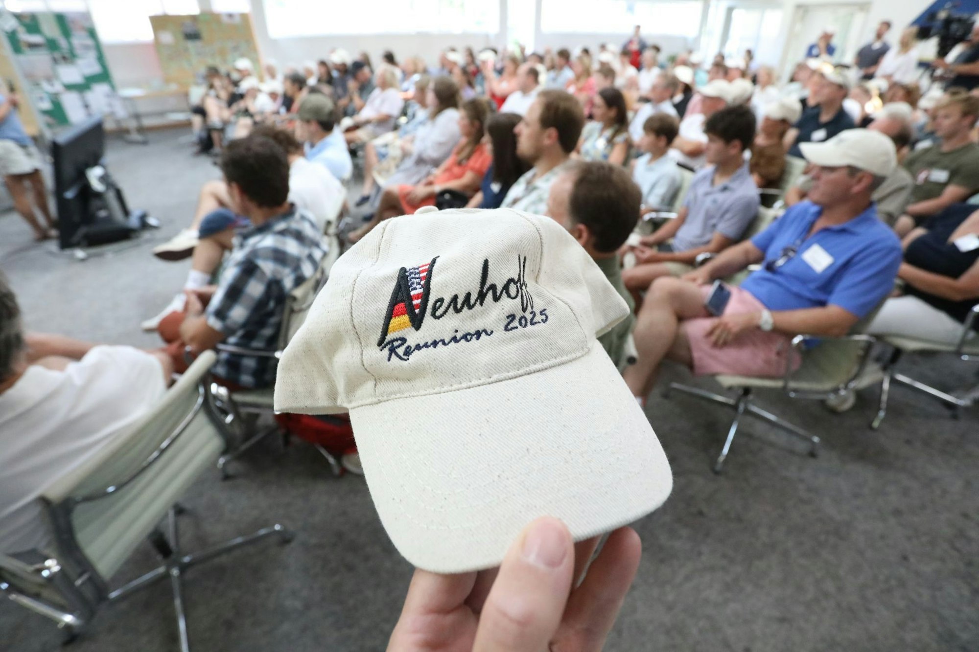 Eine Basecap mit der Aufschrift „Neuhoff – Reunion 2025“, dahinter viele Menschen, die auf Stühlen sitzen.