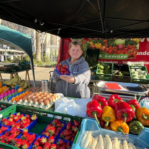 Eine Frau steht hinter ein Marktstand für Obst und Gemüse.