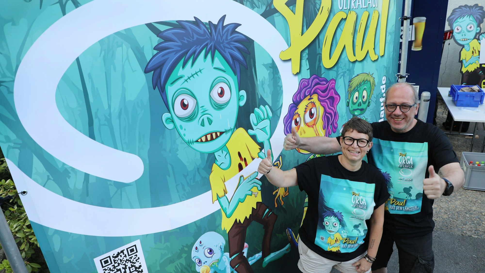 Thorsten Klenke aus Lindlar und Katja Dasbach aus Stebach bei Montabaur vor dem Dorfgemeinschaftshaus in Lindlar-Remshagen stehen vor einer Plakatwand des Panoramasteig-Ultralaufs „Paul“.