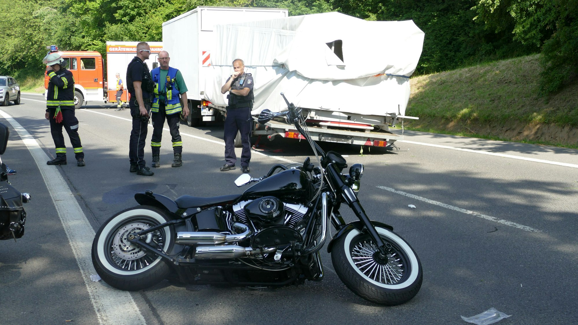 Ein schwarzes Motorrad liegt auf einer Straße auf der Seite. Im Hintergrund steht ein beschädigter Lkw.