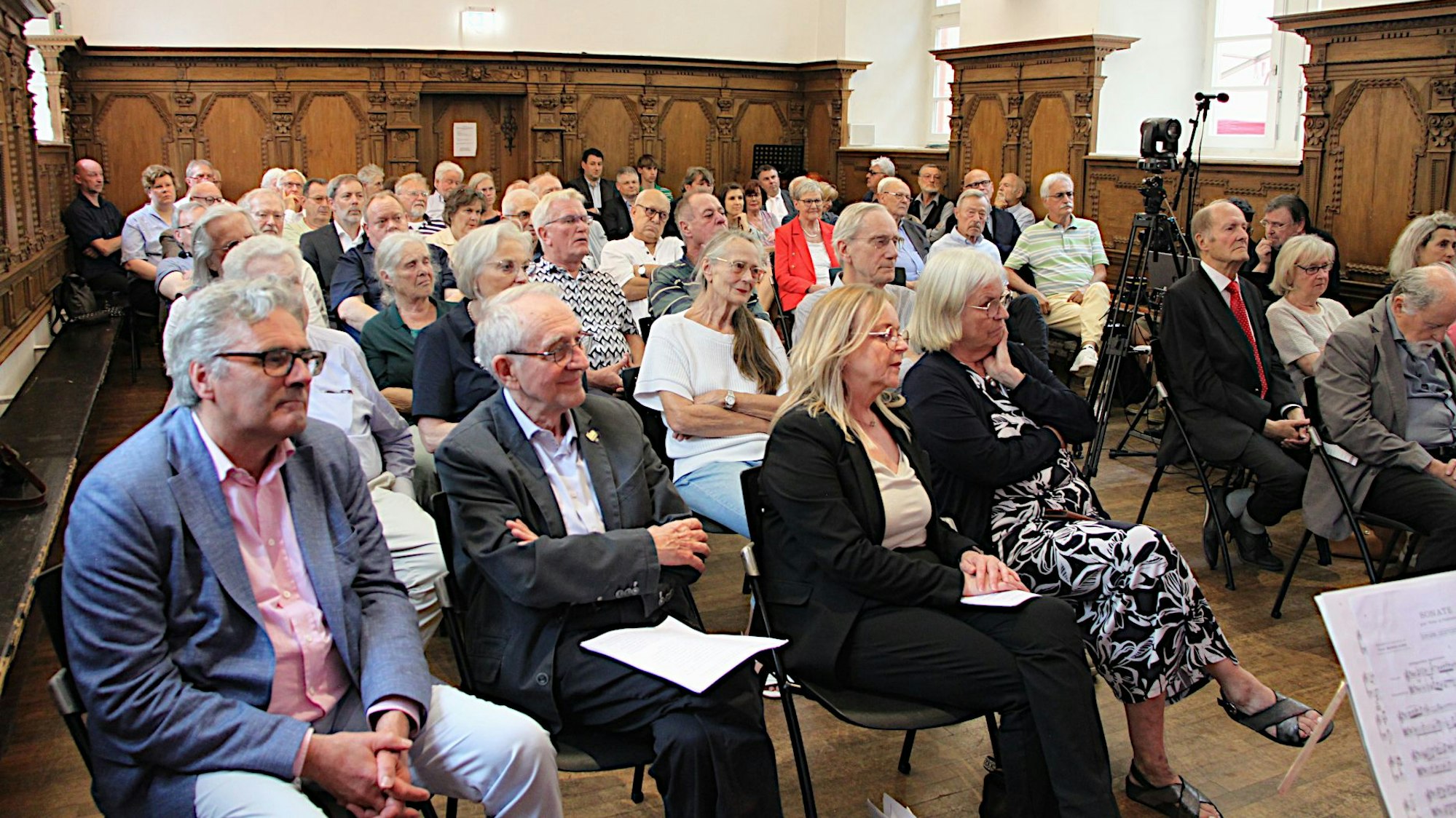 Das Bild zeigt die Teilnehmerinnen und Teilnehmer während der Feier in einem Raum mit Holzvertäfelung an den Wänden.