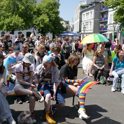 Menschen sitzen in Regenbogenfarben und mit Pride-Flaggen auf Bänken am Siegburger Markt.