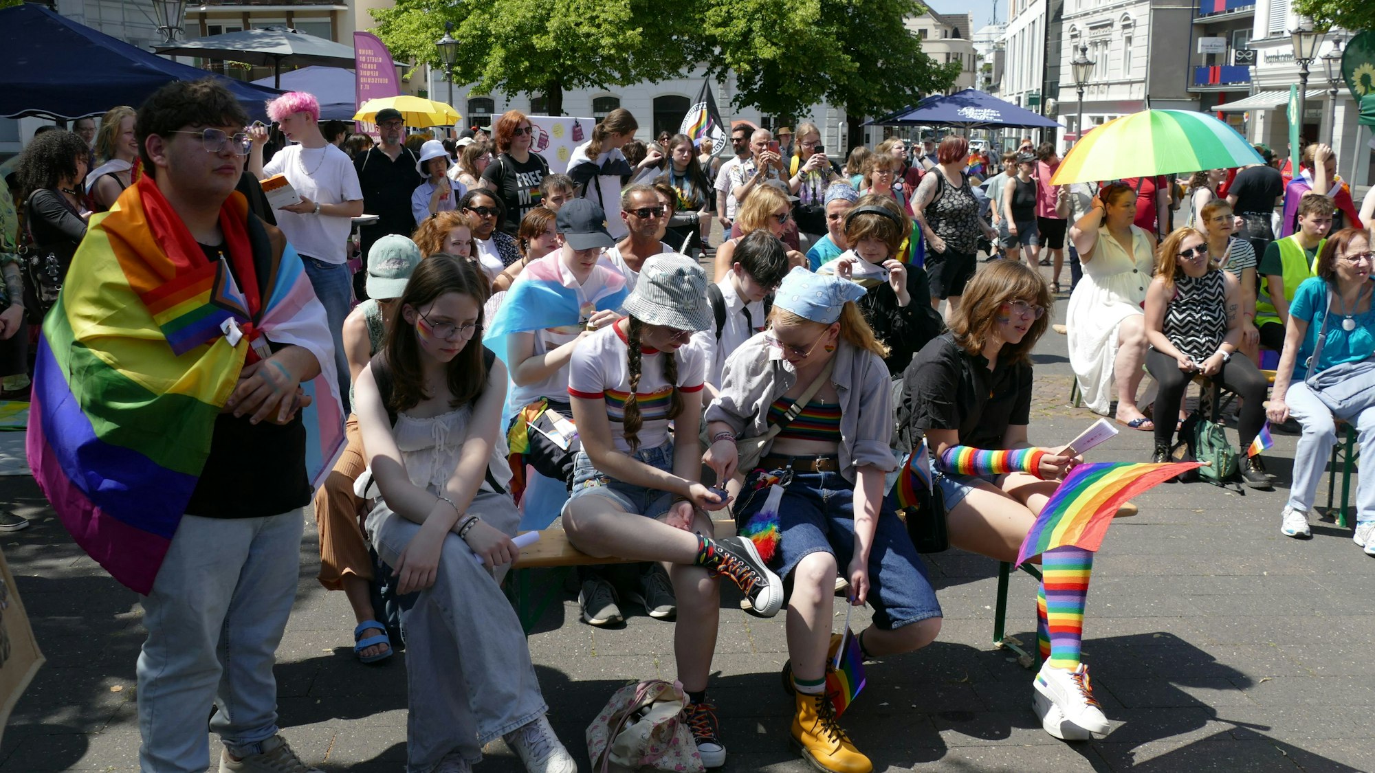 Menschen sitzen in Regenbogenfarben und mit Pride-Flaggen auf Bänken am Siegburger Markt.
