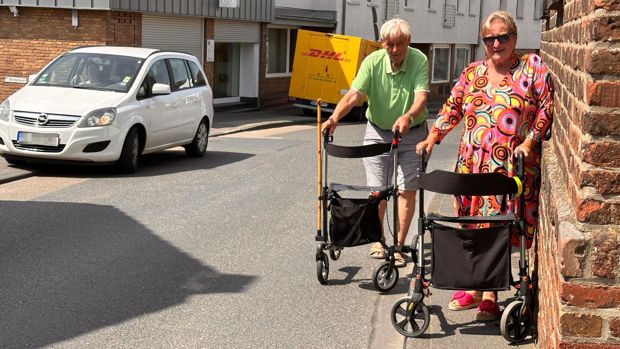 An einer Straße versuchen zwei Frauen mit Rollatoren auf einem viel zu schmalen Gehweg zu bleiben.