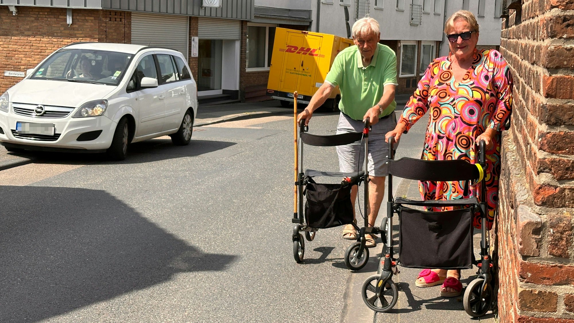 An einer Straße versuchen zwei Frauen mit Rollatoren auf einem viel zu schmalen Gehweg zu bleiben.