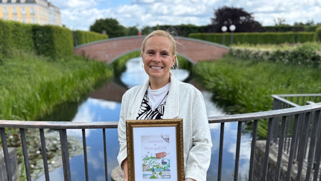 Auf dem Bild ist eine Frau im Schlosspark Brühl zu sehen, die einen Bildrahmen mit dem Buchtitel in der Hand hält.