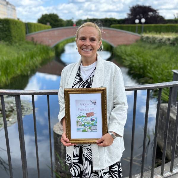 Auf dem Bild ist eine Frau im Schlosspark Brühl zu sehen, die einen Bildrahmen mit dem Buchtitel in der Hand hält.