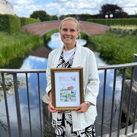 Auf dem Bild ist eine Frau im Schlosspark Brühl zu sehen, die einen Bildrahmen mit dem Buchtitel in der Hand hält.