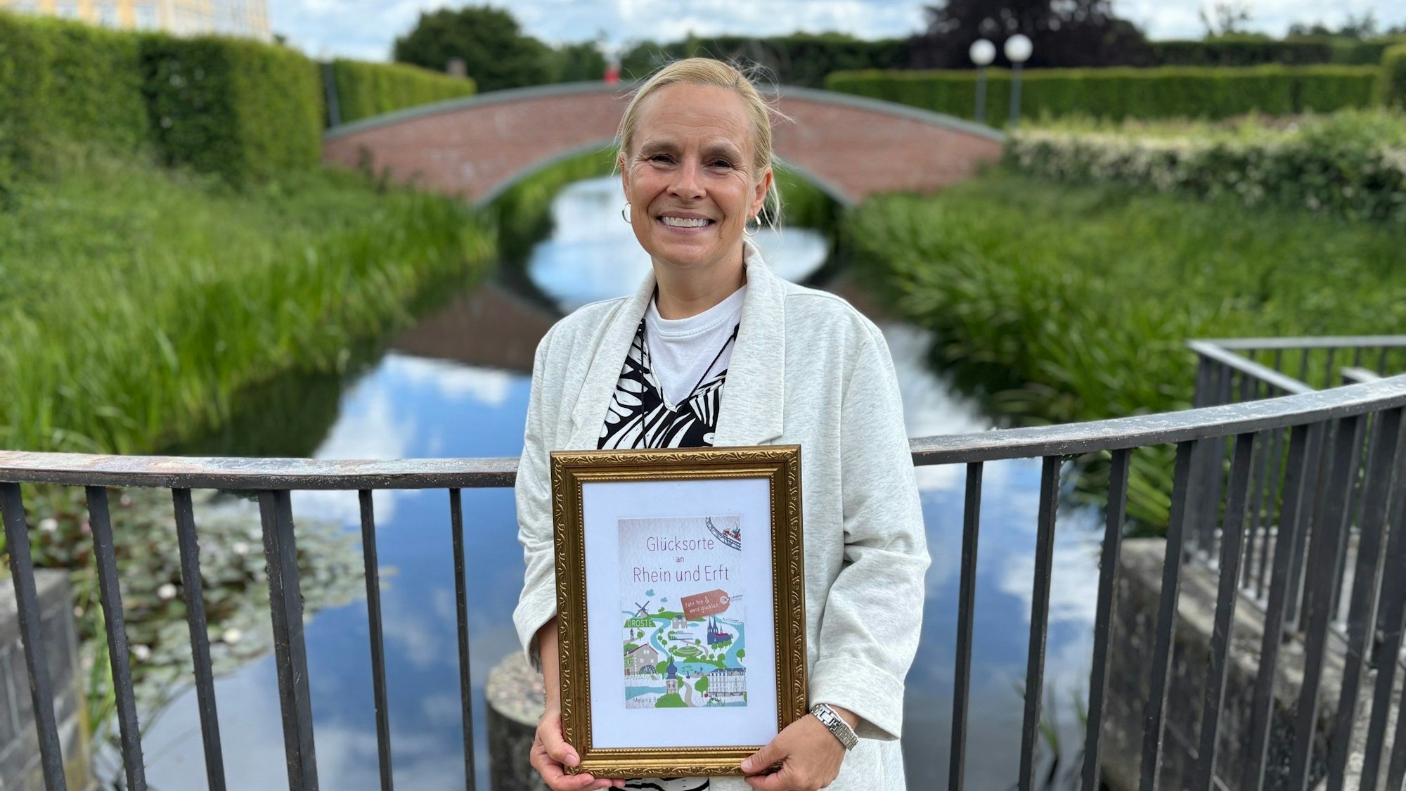 Auf dem Bild ist eine Frau im Schlosspark Brühl zu sehen, die einen Bildrahmen mit dem Buchtitel in der Hand hält.