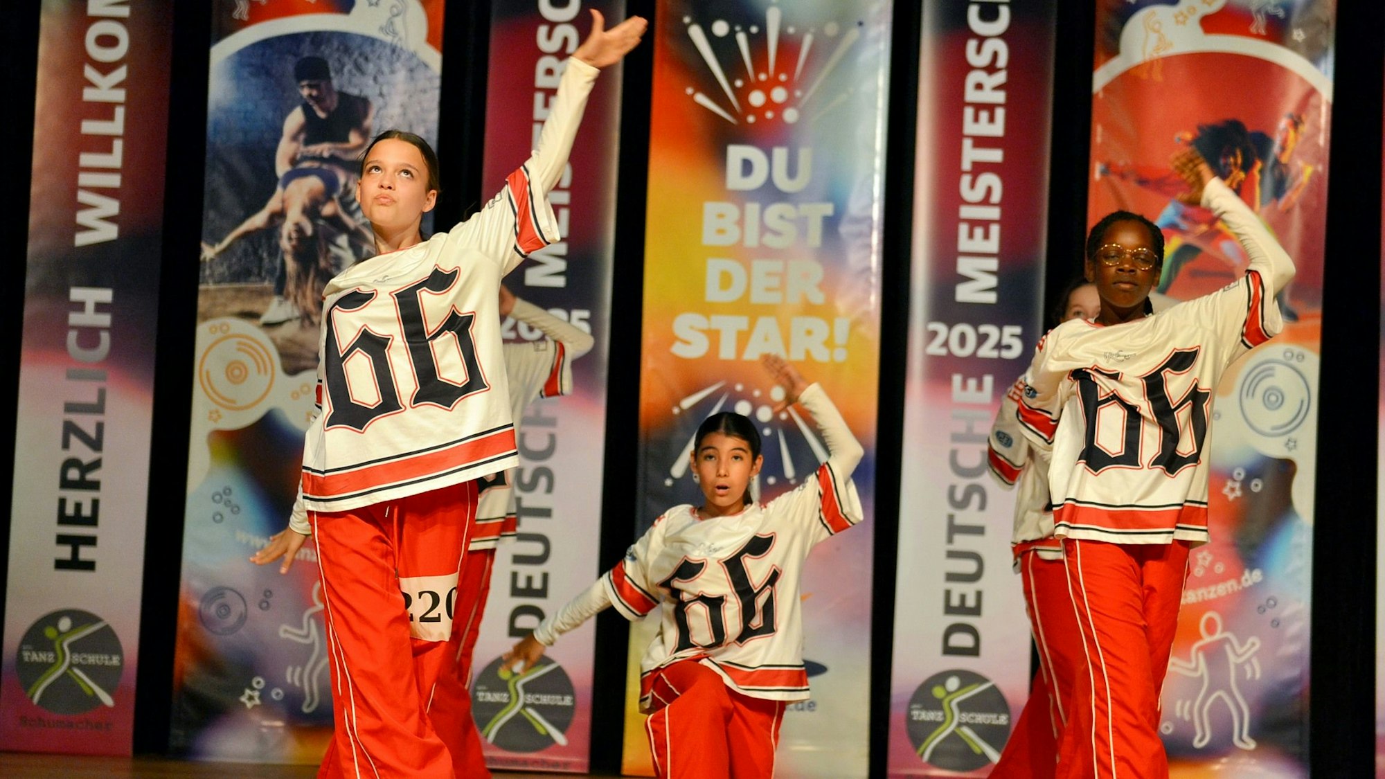 Ein Tanztrio bestehend aus zwei Mädchen und einem Jungen, der hinten in der Mitte steht, sind auf der Bühne. Sie tragen rote Hosen und Shirts mit der Aufschrift "66". Alle haben den linken Arm in die Höhe gereckt. Die Bewegungen sind nicht synchron.