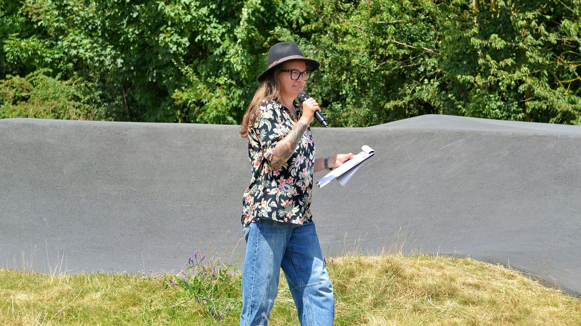 Eine Frau mit Hut, geblümter Bluse und Jeans steht am Pumptrack in Weilerswist und spricht in ein Mikrofon.