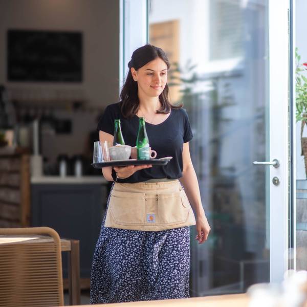 Eine junge Frau läuft mit einem Tablett, auf dem Kaffeetassen und Wasserflaschen stehen, zu einem Tisch.