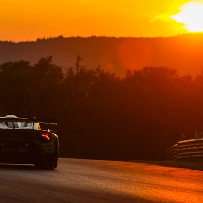 Das Bild zeigt einen Rennwagen, der auf dem Nürburgring fährt. Im Hintergrund geht die Sonne unter.