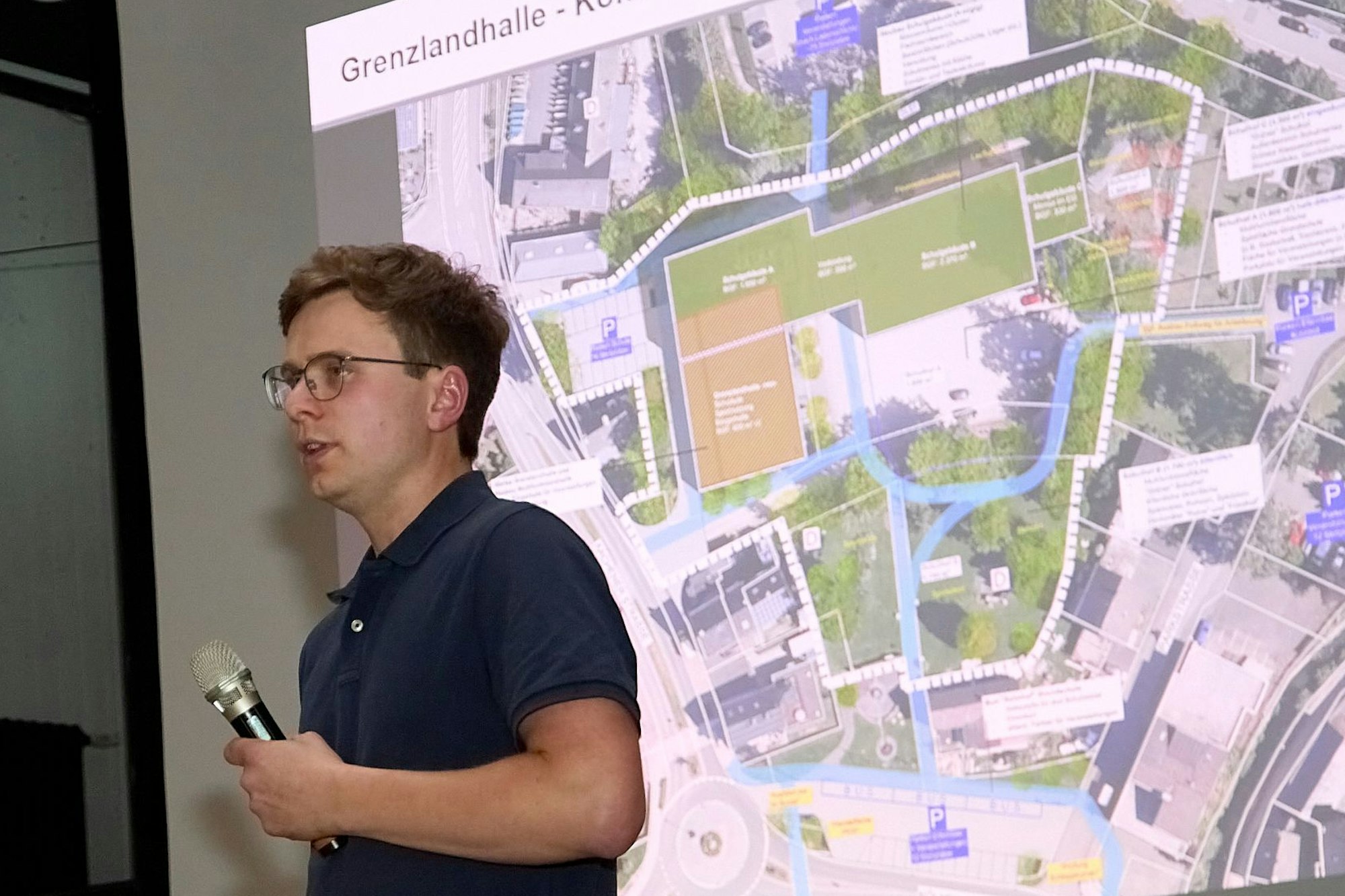 Christian Schmitz hält ein Mikrofon in der Hand. Hinter ihm ist ein Luftbild des Bereichs um die Grenzlandhalle in Hellenthal zu sehen.