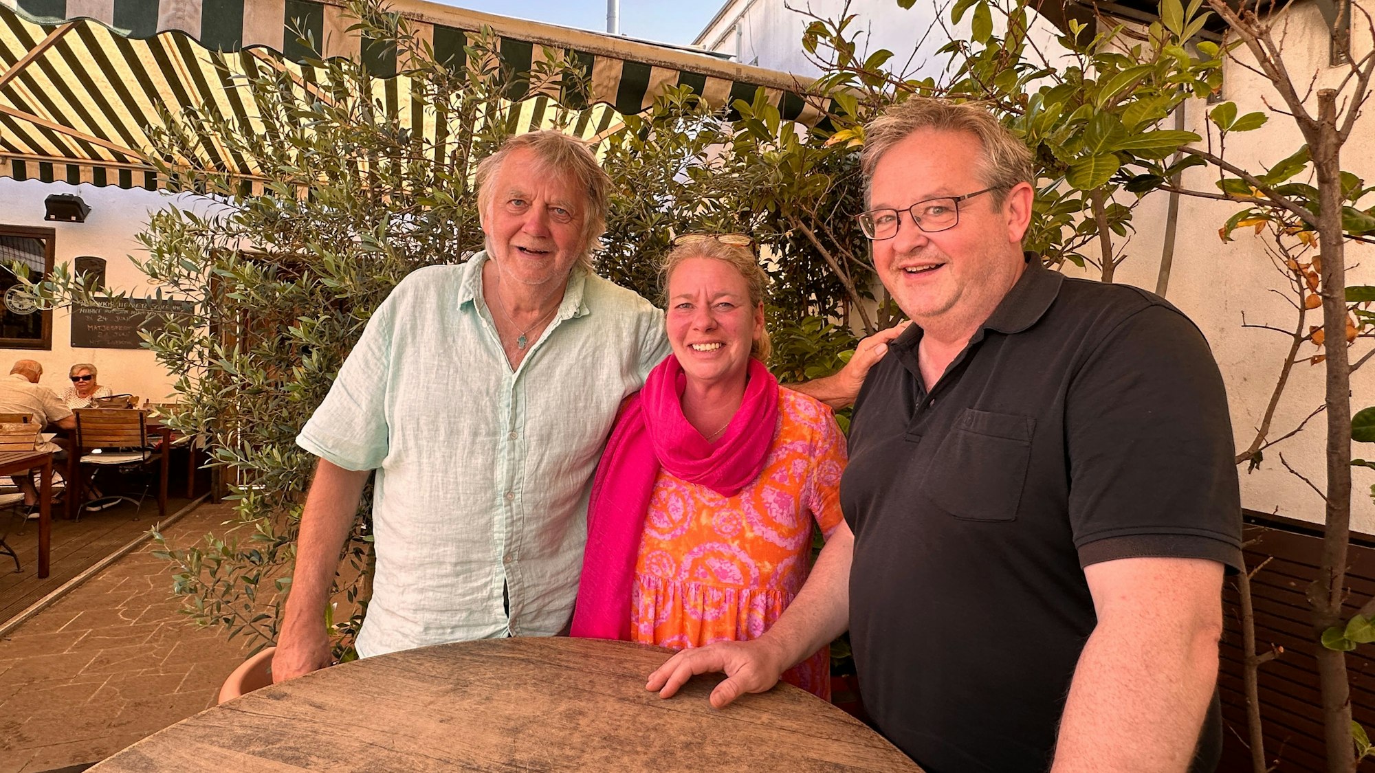 Zwei Männer und eine Frau stehen an einem Stehtisch in einem Biergarten.