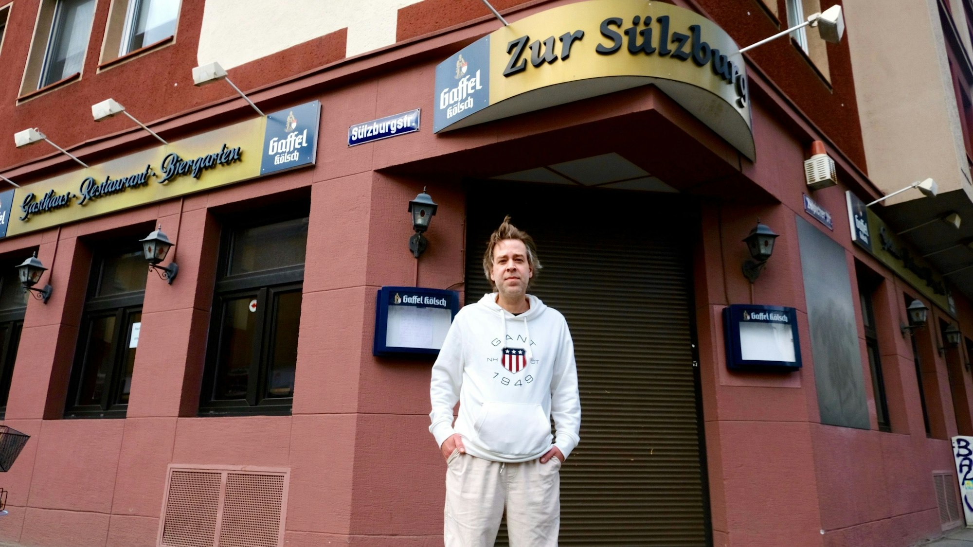 Ein Mann steht vor einer Eckkneipe mit heruntergelassenen Rollläden.