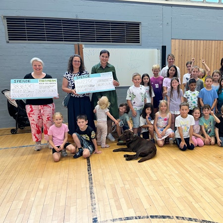 Viele Kinder hocken in einer Turnhalle, ein Hund ist auch dabei. Vier Frauen halten großformatige Spenden-Schecks in die Höhe.