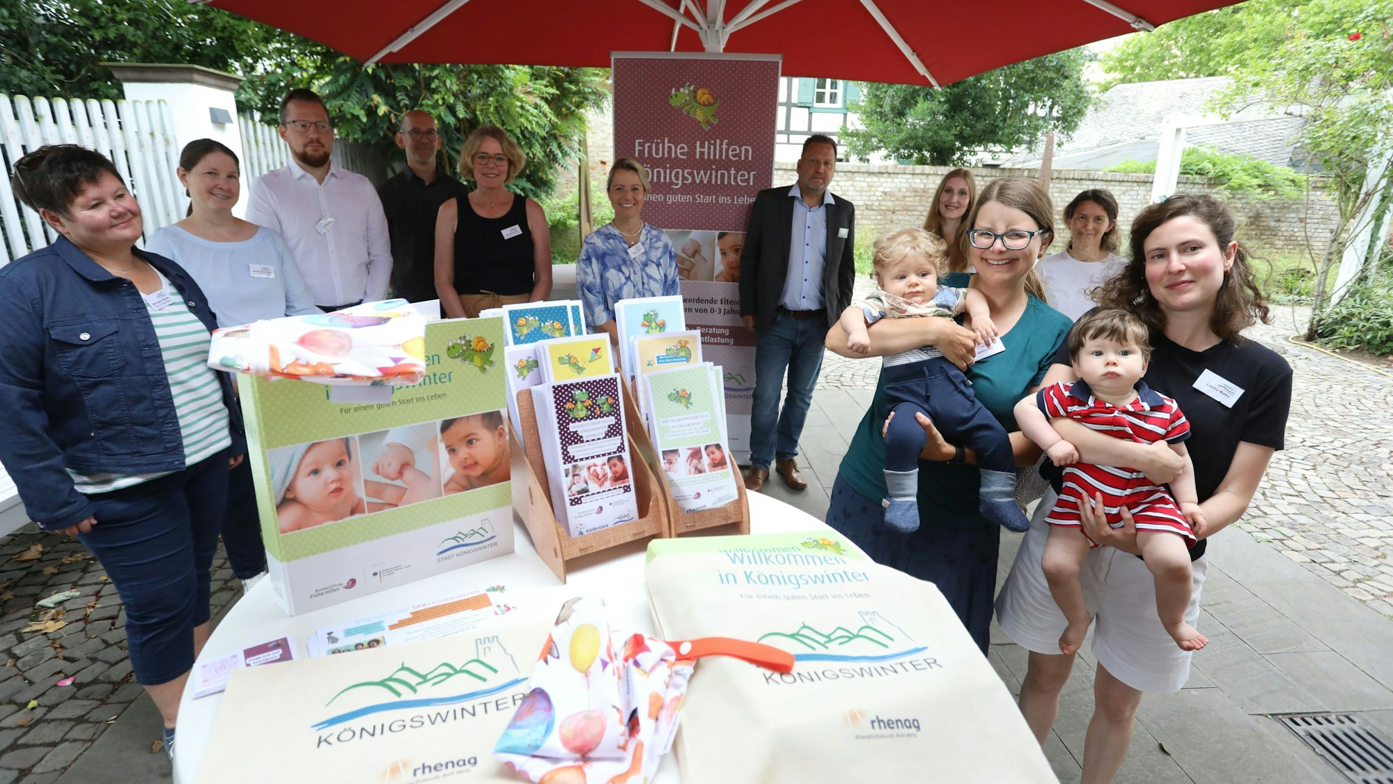 Eine Gruppe von Menschen hinter einem Stehtisch mit Info-Material. Zwei Mütter halten ihre Babys auf dem Arm.