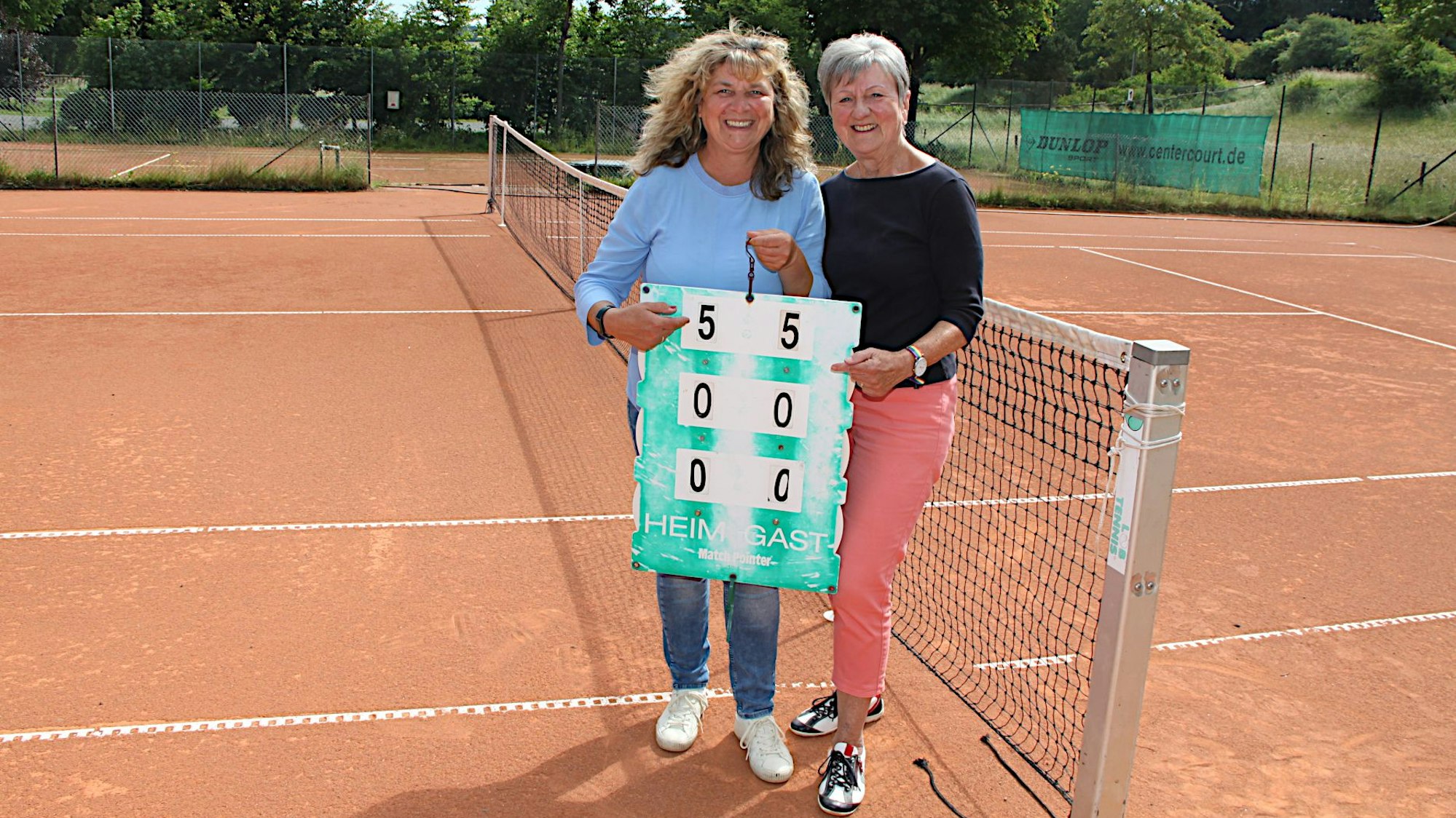 Zwe Frauen stehen auf einem Tennisplatz und halten ein Schild hoch.