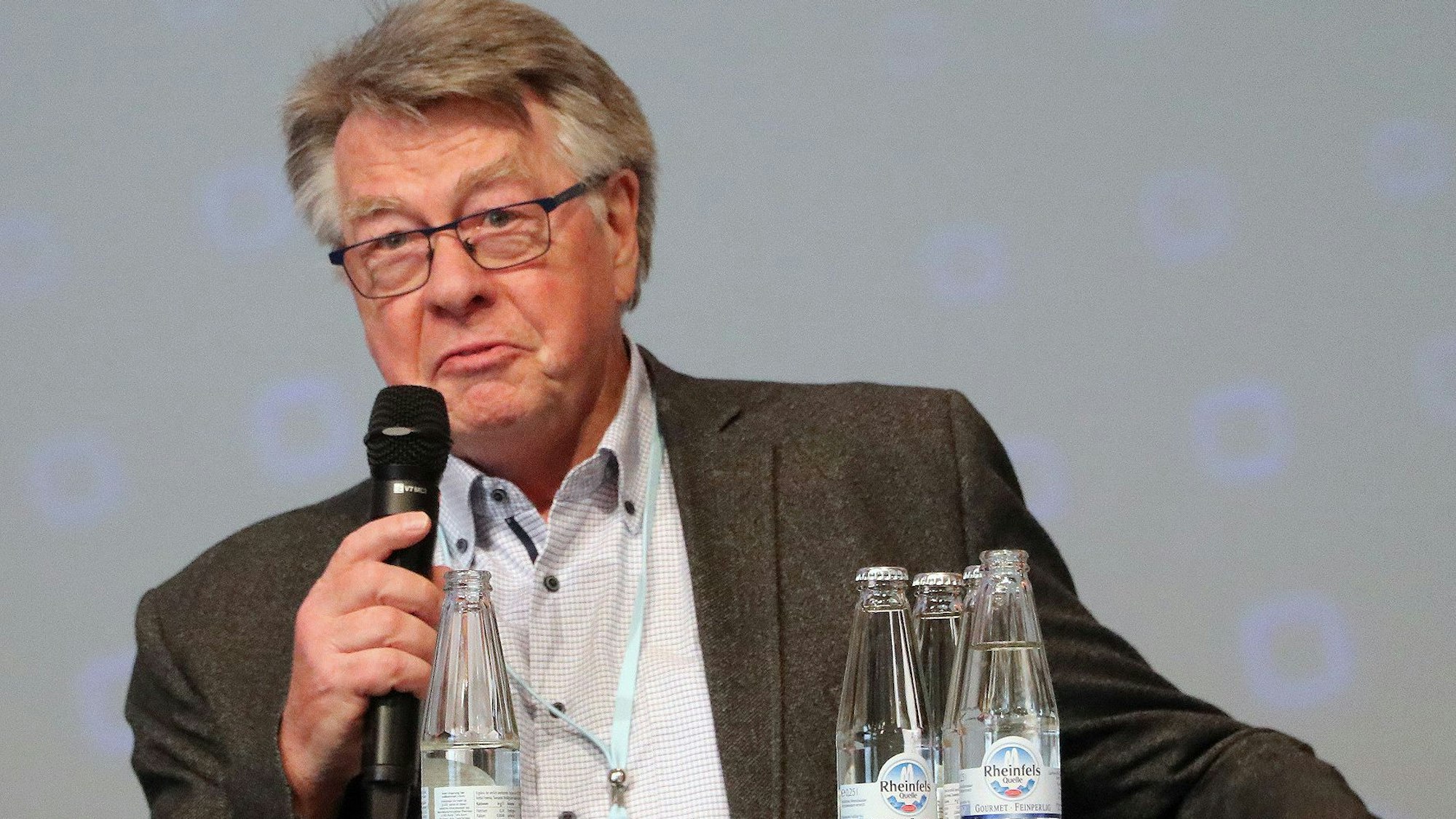 Auf dem Foto ist ein Mann mit Brille und grauem Haar zu sehen. Er hält ein Mikrofon in seiner Hand.
