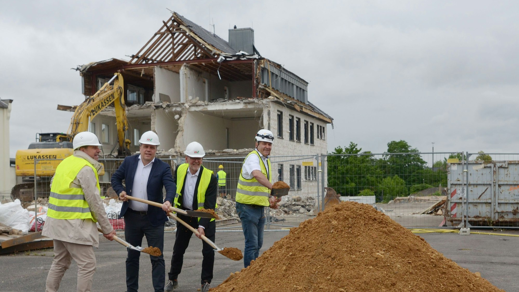 Vier Männer mit Schutzhelmen werfen mit Spaten Erde in die Luft. Im Hintergrund bricht ein Bagger ein Gebäude ab.