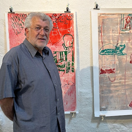 Ein Mann mit grauem Kurzhaarschnitt, Dreitagebart und Brille steht vor einer Wand, an der zwei Drucke hängen.