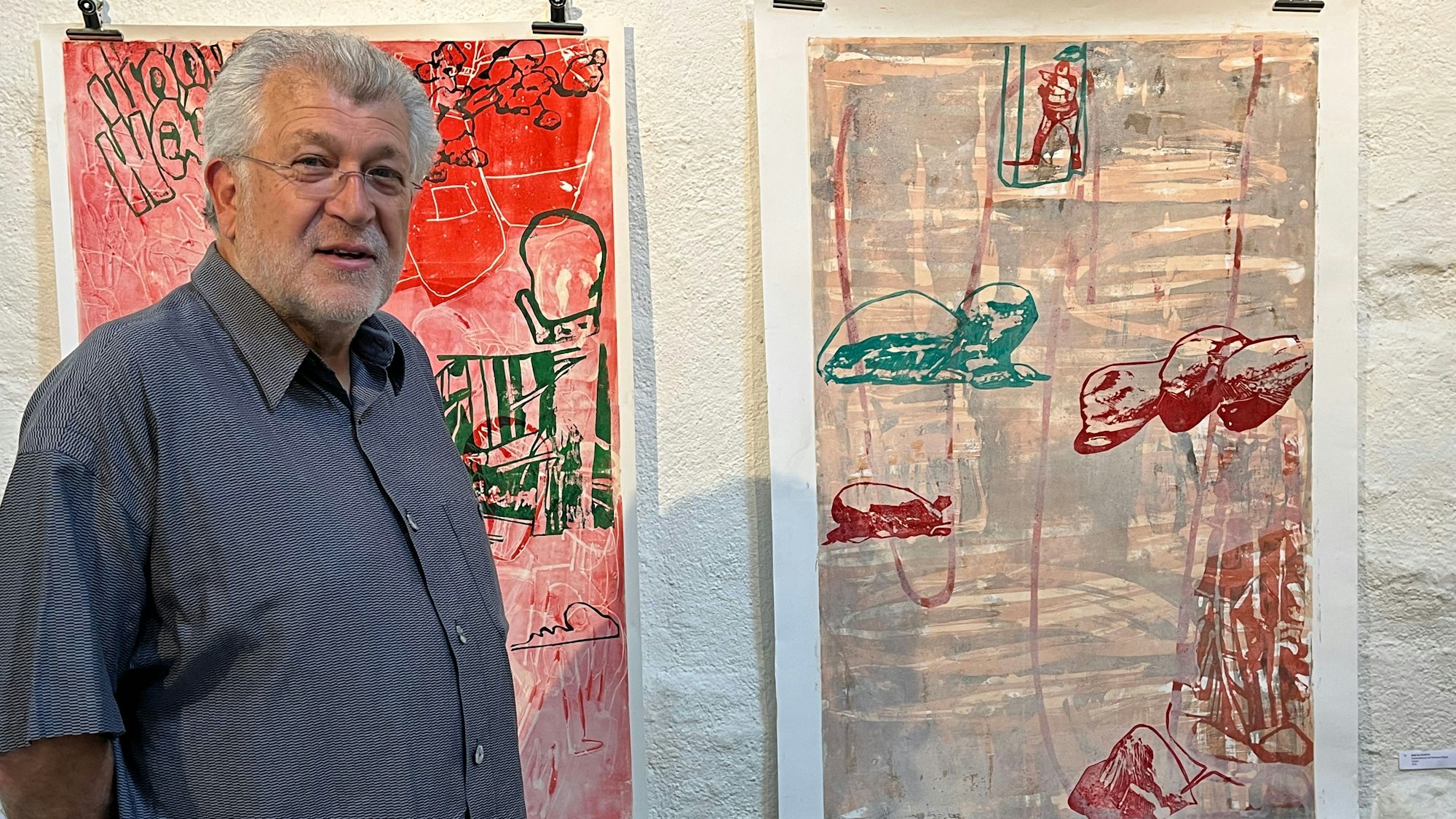 Ein Mann mit grauem Kurzhaarschnitt, Dreitagebart und Brille steht vor einer Wand, an der zwei Drucke hängen.