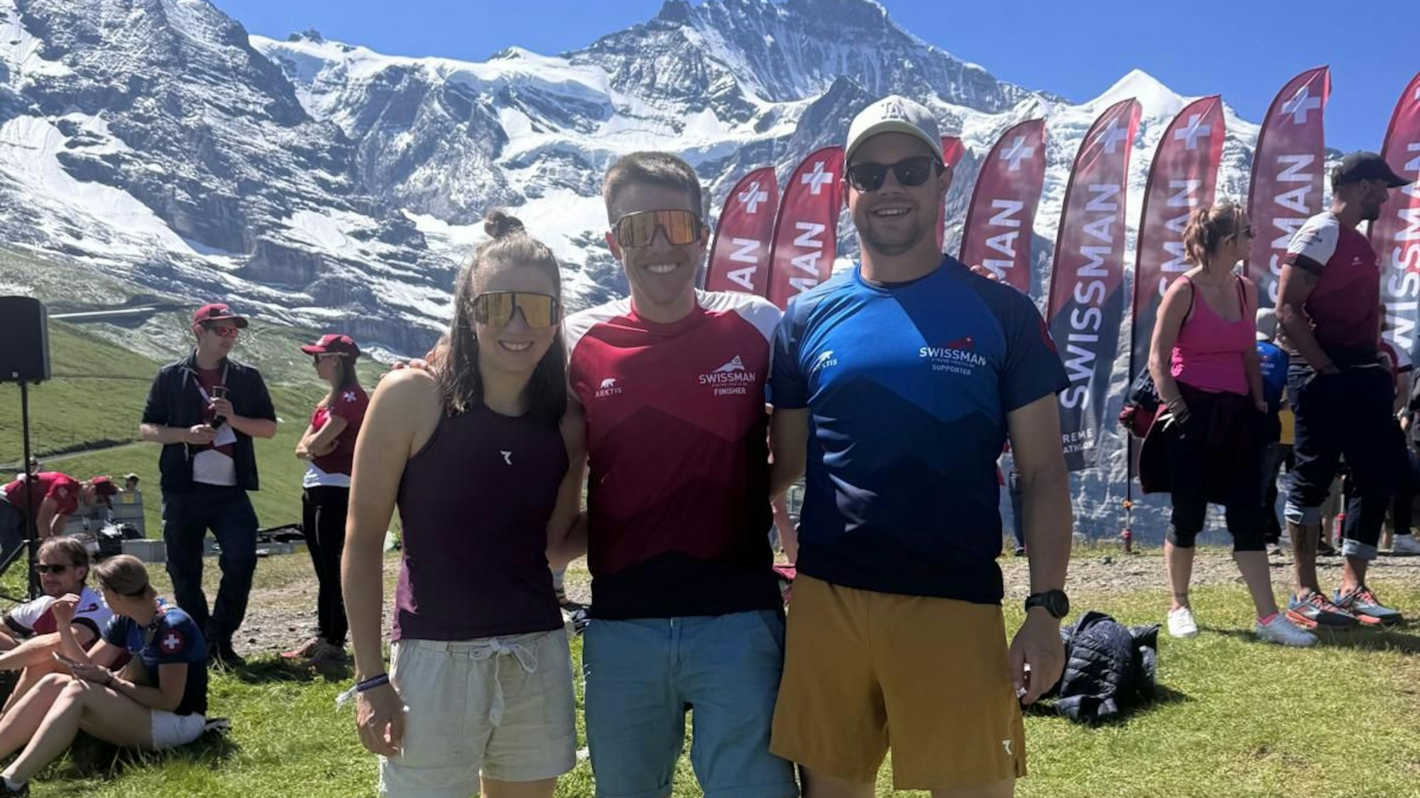 Zwei Männer und eine Frau stehen nach absolviertem Triathlon vor einer Bergkulisse in der Schweiz.