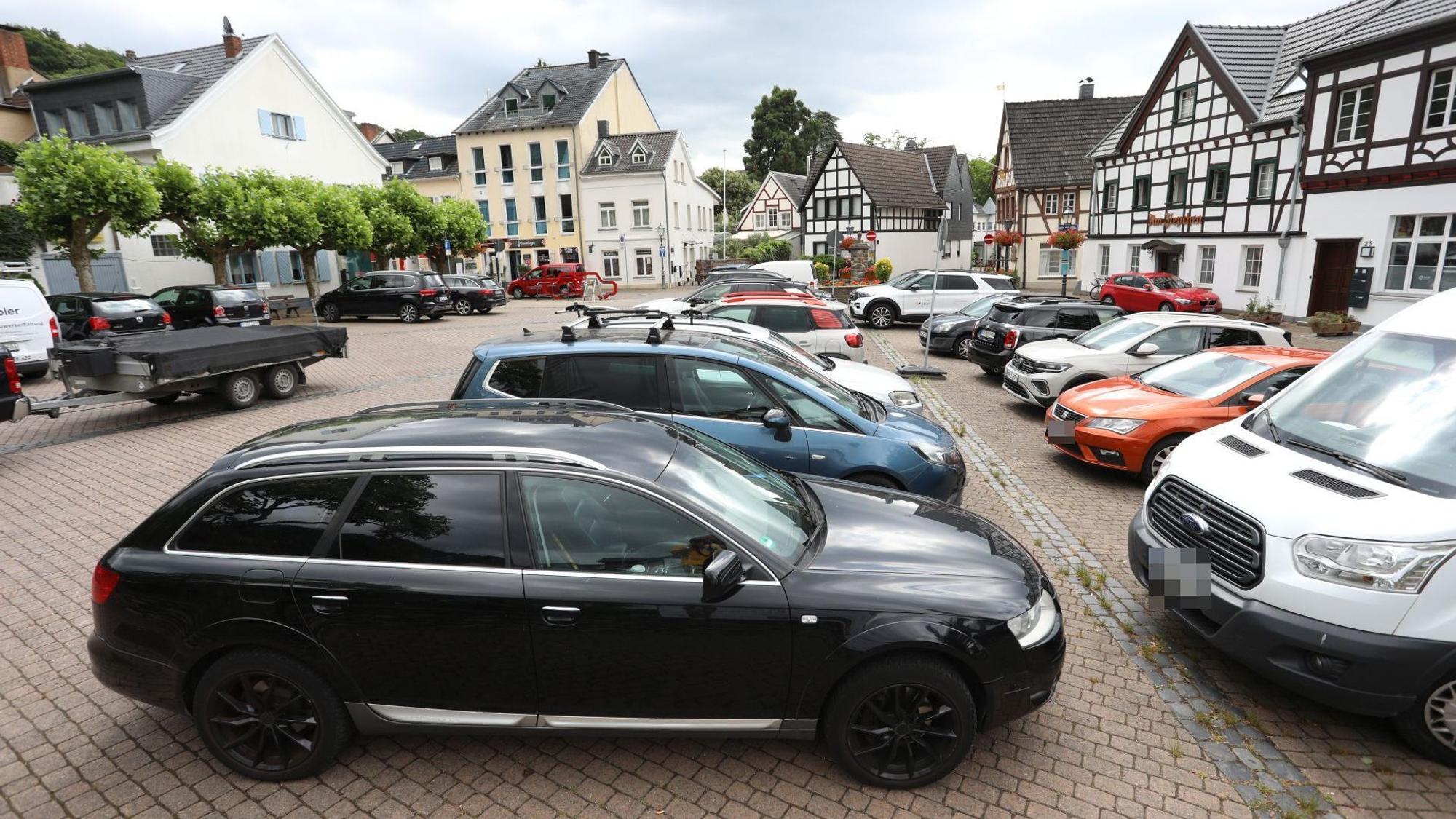 Autos stehen auf einem Dorfplatz, im Hintergrund alte Fachwerkhäuser.