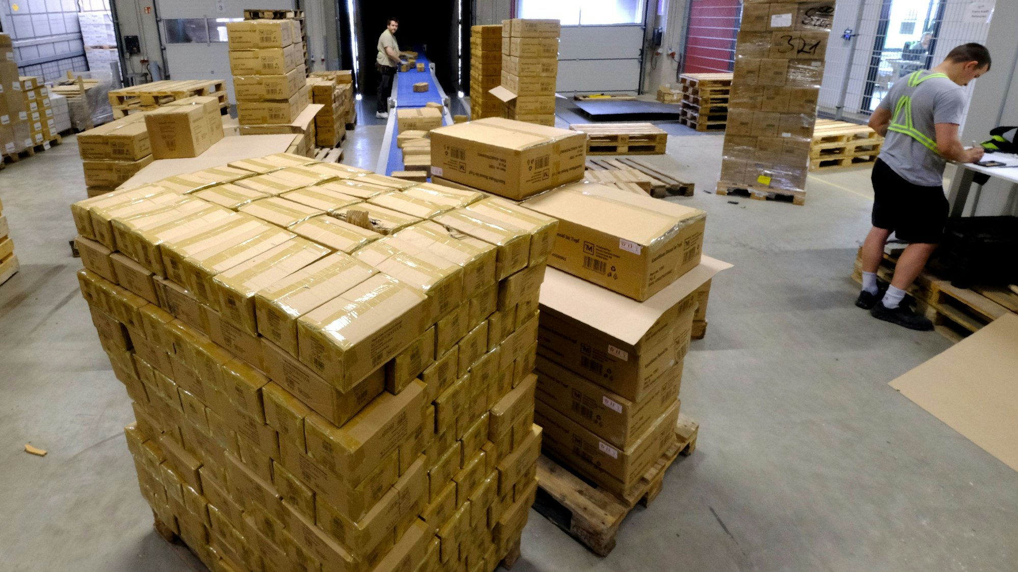 Über ein Transportband werden Pakete aus einem Container entladen und von Mitarbeitern auf einer Palette gestapelt.
