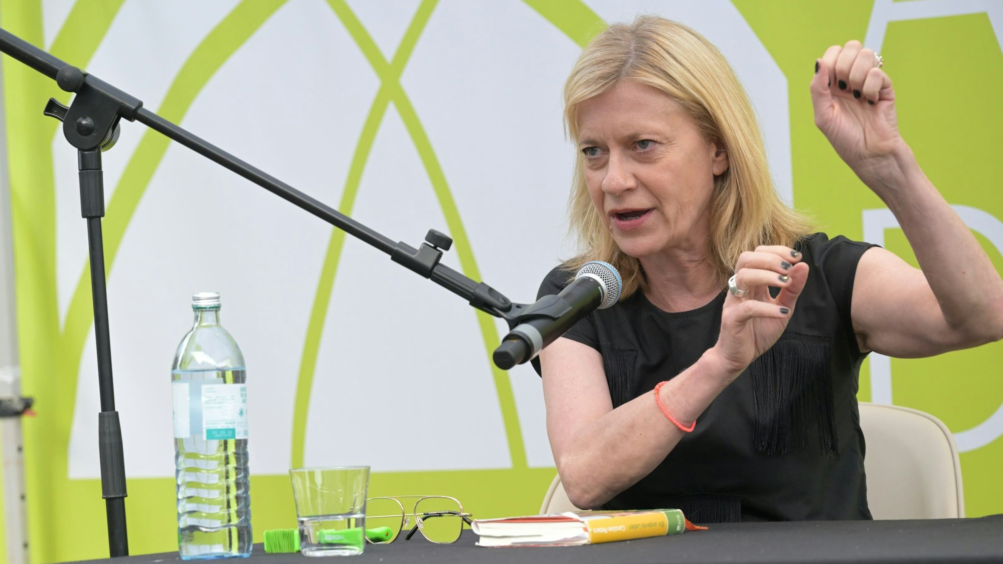 Caroline Peters spricht in ein Mikrofon, vor ihr liegt ihr Buch, eine Brille und eine Wasserflasche.
