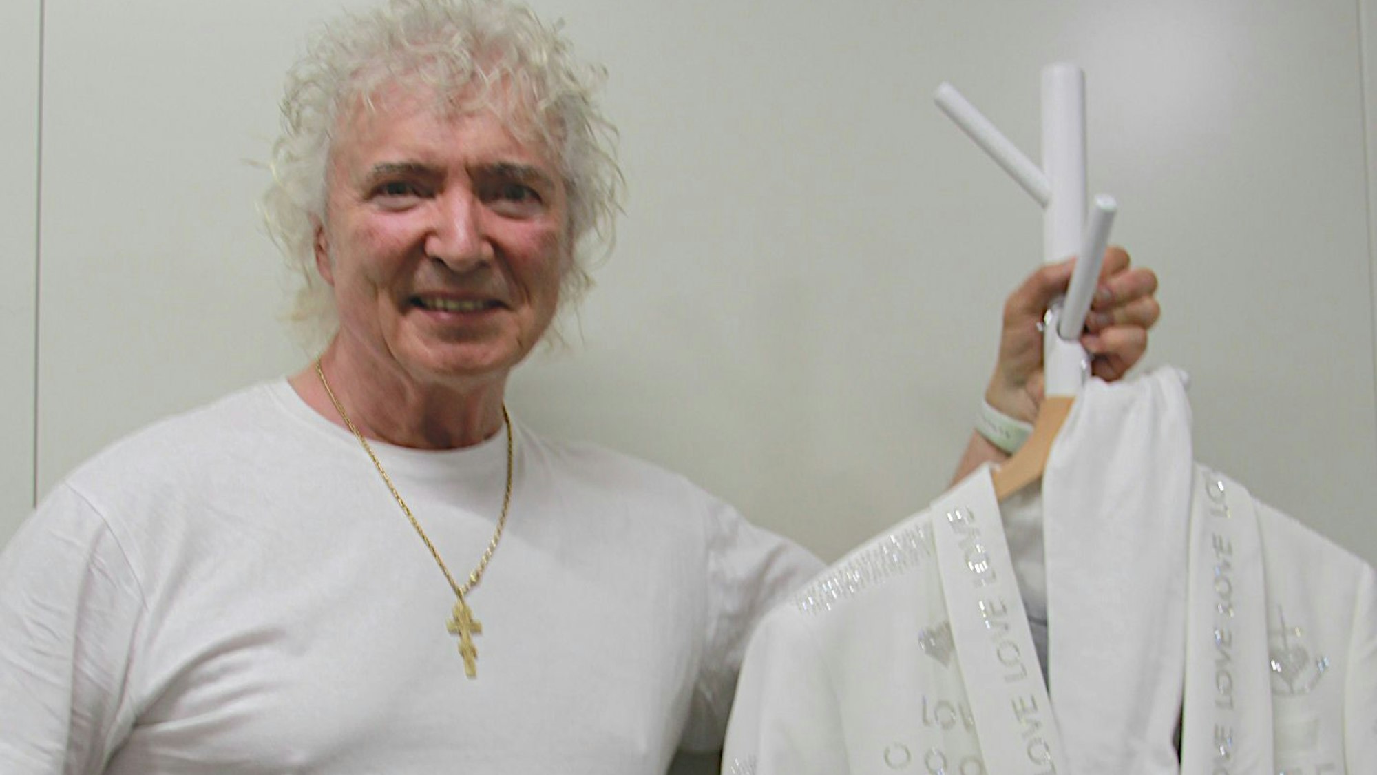 Peter Orloff steht in einem weißen T-Shirt neben einem Kleiderständer, an dem ein weißer Anzug hängt.