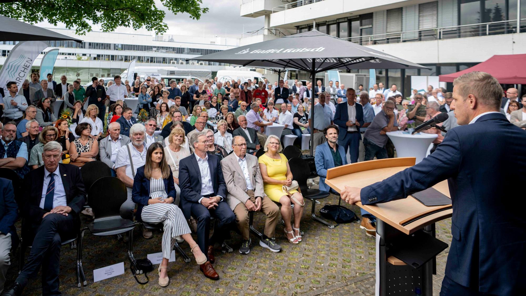 Markus Ramers, Landrat im Kreis Euskirchen, steht an einem Pult und spricht in ein Mikrofon. Vor ihm sitzen zahlreiche Menschen und hören zu.
