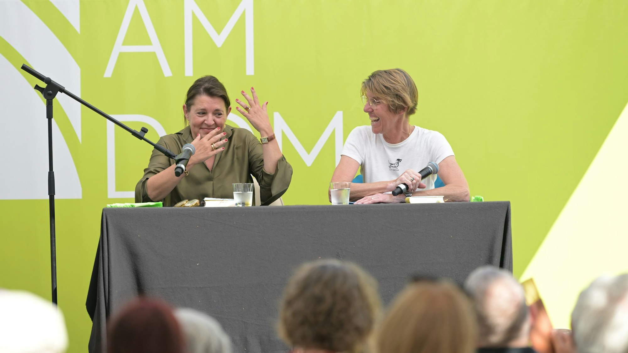 Zwei Frauen sitzen auf einer Bühne, hinter ihnen eine grasgrüne Wand.