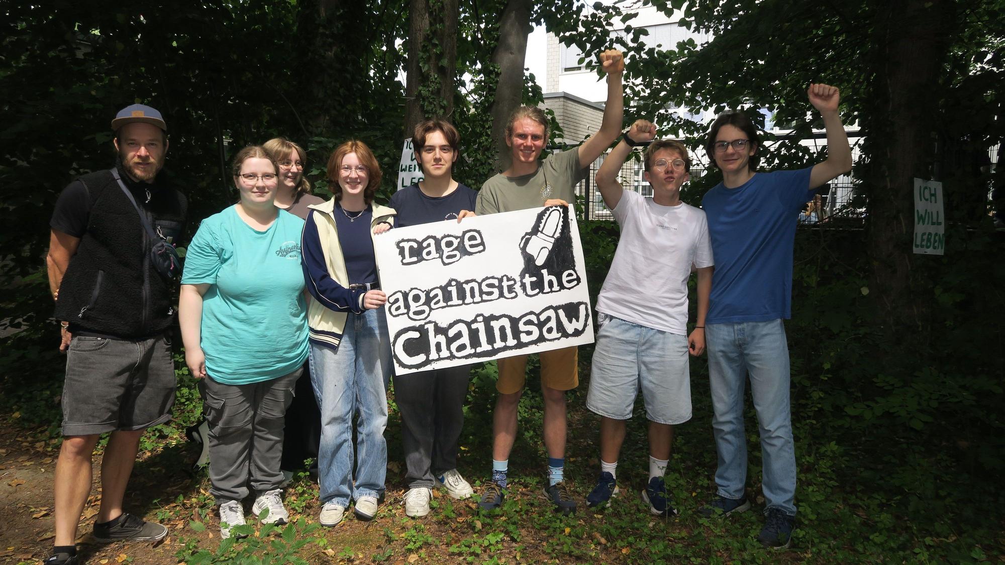 Eine Schülergruppe steht unter Bäumen und hält ein Plakat hoch mit der Aufschrift „Rage against the chainsaw“.