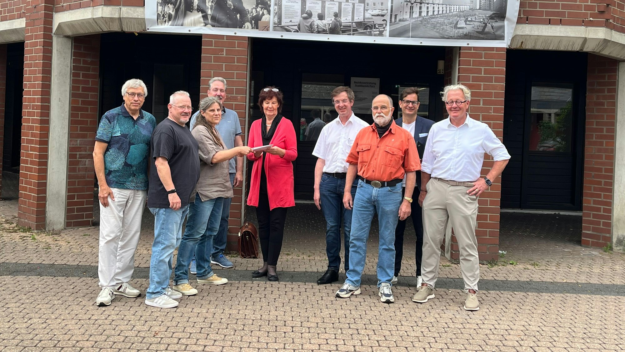 Eine Frau übergibt der Porzer Bezirksbürgermeisterin eine Unterschriftenliste vor dem Bezirksrathaus Porz. Um die beiden Frauen herum stehen sieben Männer.