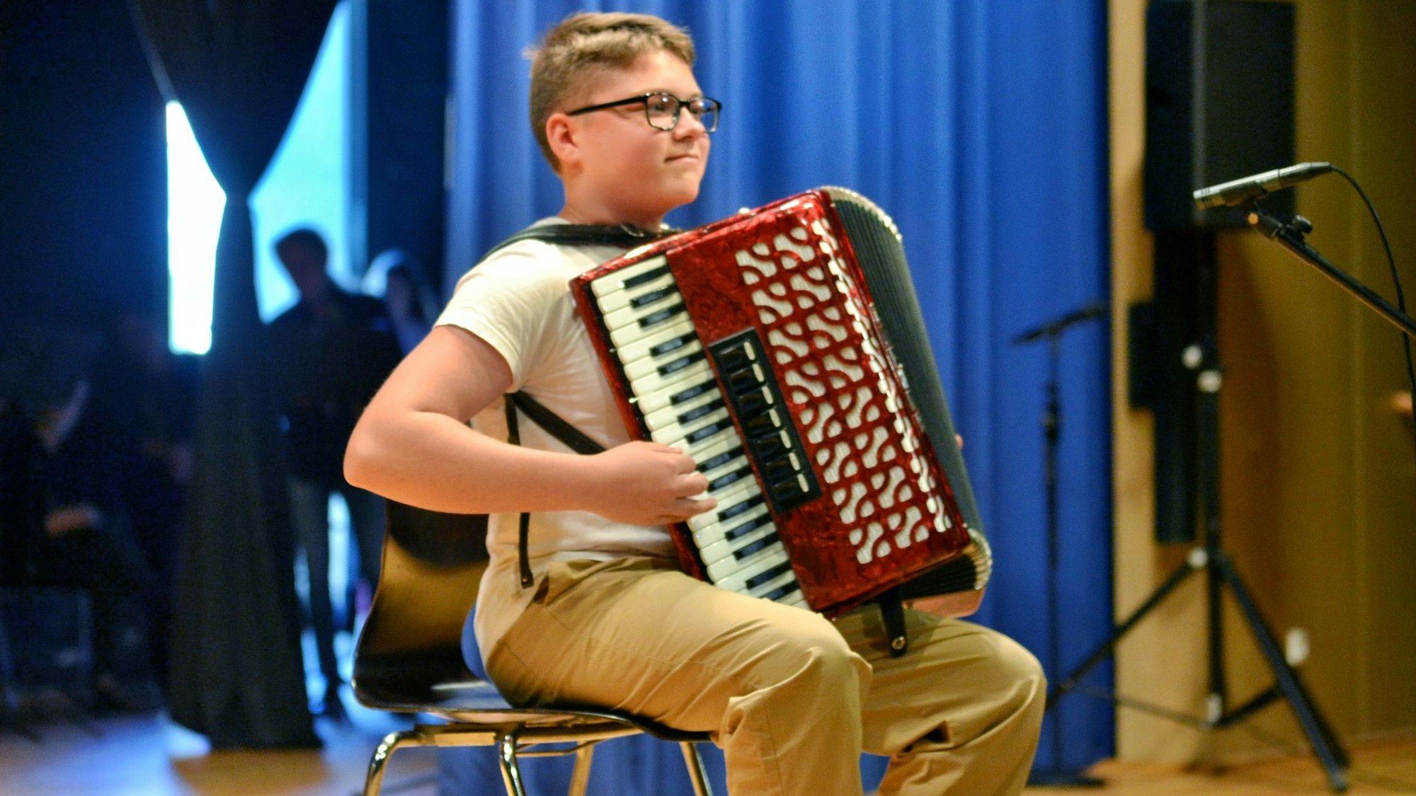 Ein Junge mit weißem T-Shirt und beiger Hose sitzt auf einem Stuhl und spielt Akkordeon.