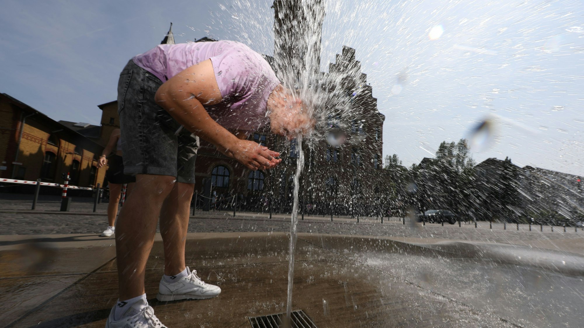 Ein Mann hält seinen Kopf in den Wasserstrahl eines Brunnens, der aus dem Boden kommt.